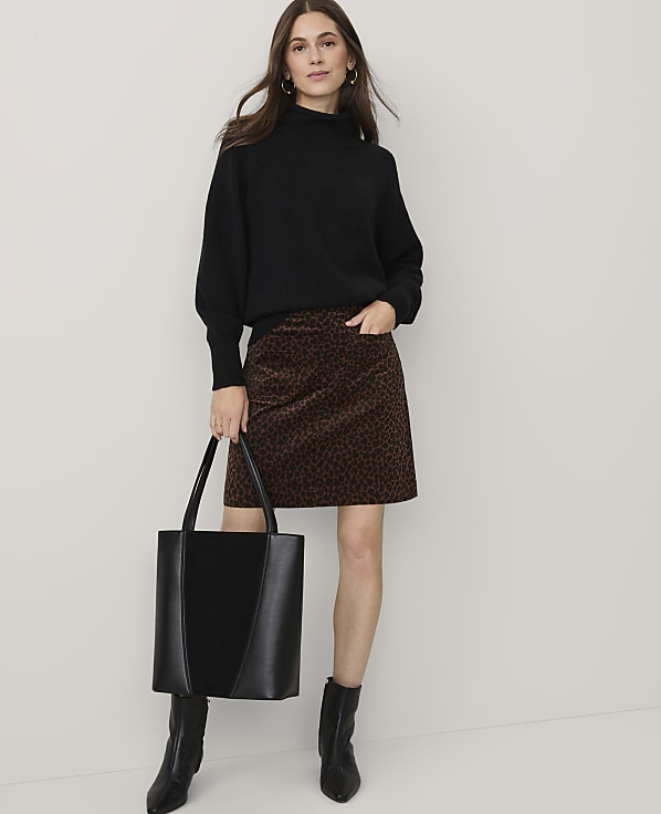 A woman wearing a black sweater, leopard-print skirt, and black boots stands holding a black tote bag against a plain white background.