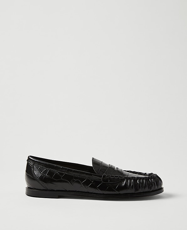 A single black crocodile-embossed leather loafer sits in the foreground against a plain, light grey background.