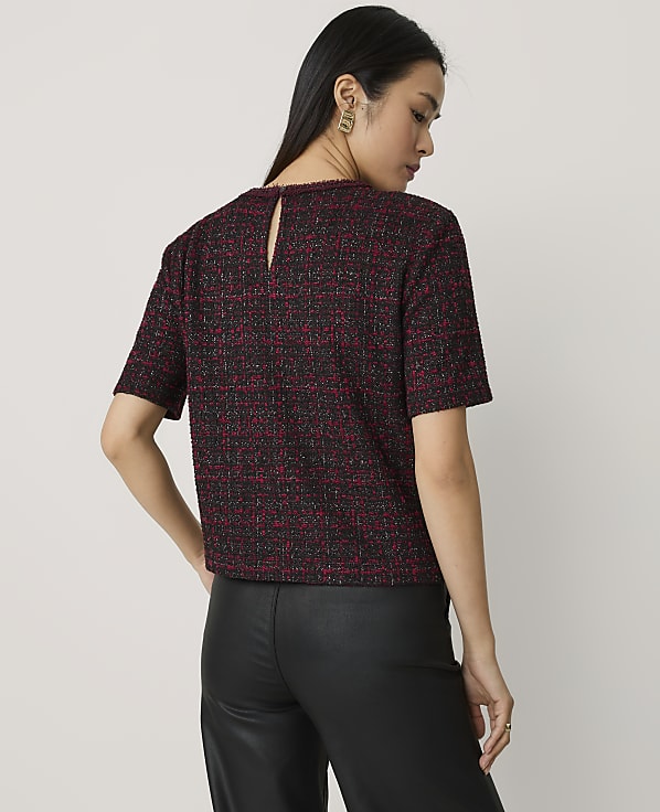 A woman wearing a red and black tweed short-sleeved top and black leather trousers stands against a plain light grey background.