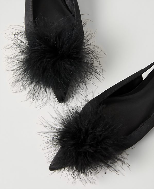 Two black satin pointed-toe shoes with fluffy black feather pom-poms are set against a plain white background.