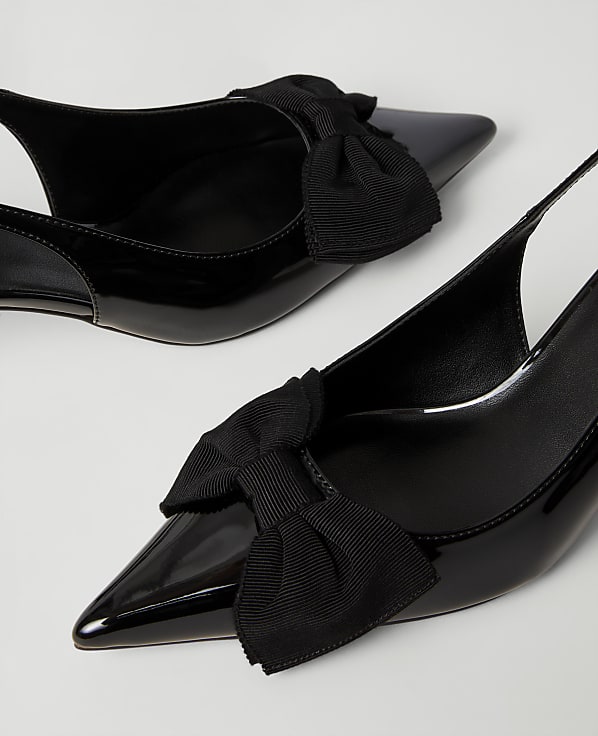 Two black patent leather pointed-toe heels with decorative bows sit against a plain white background.
