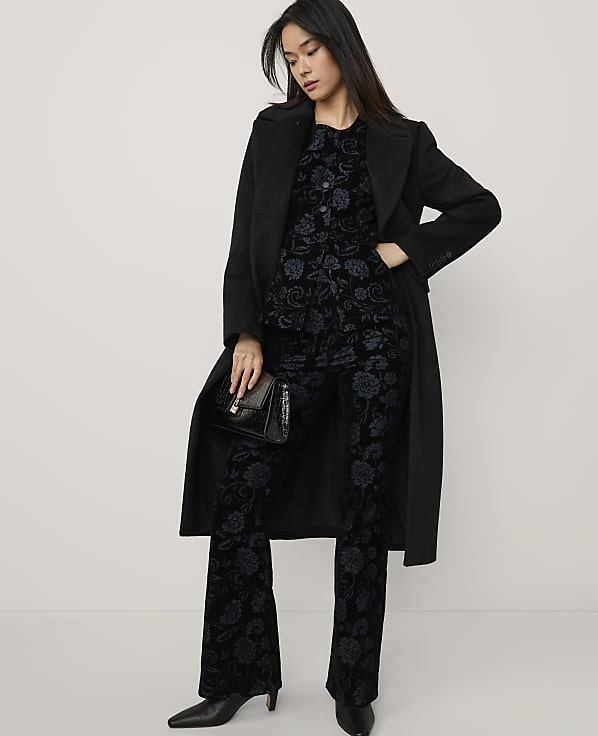 A woman wearing a black coat over a dark floral patterned suit and holding a clutch stands against a plain, light-colored background.