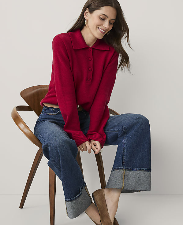A woman wearing a red collared sweater and wide-leg cuffed jeans sits in a wooden chair against a plain white background.