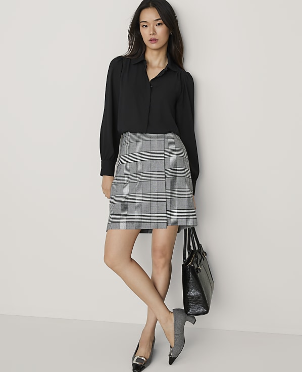 A woman wearing a black blouse and a grey plaid skirt stands against a plain white wall while holding a black handbag.