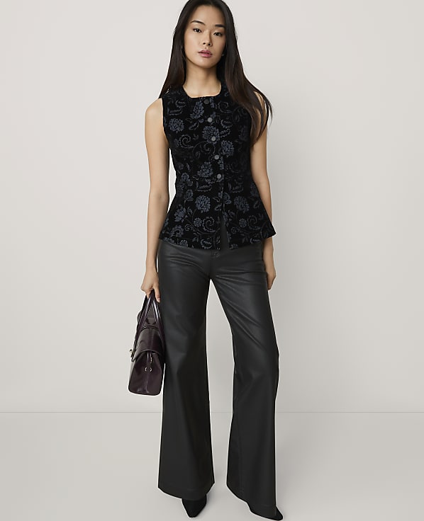 A woman wearing a black patterned vest and black leather flared trousers stands against a plain, light-colored background.