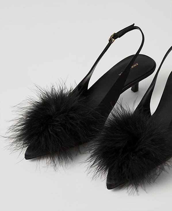 A pair of black pointed-toe heels with large fluffy pom-poms on the front are set against a plain white background.