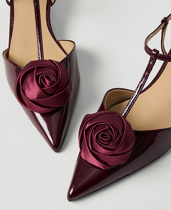 Two burgundy patent leather pointed-toe heels with fabric rose embellishments are set against a plain white background.