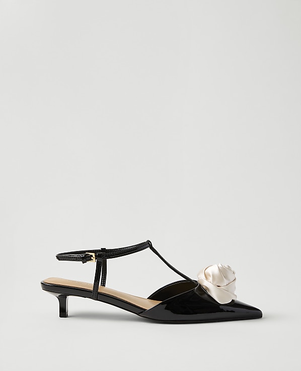 A black patent leather kitten heel with a cream-colored fabric flower on the toe sits against a plain, light gray background.