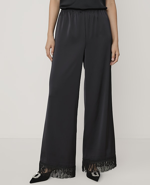 A person wears wide-leg black satin trousers with fringed hems and black pointed heels against a plain light grey background.