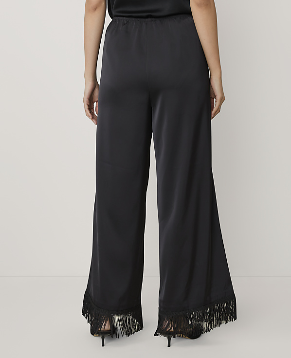 A person stands facing away from the camera wearing wide-leg black satin trousers with fringe hems against a plain light grey background.