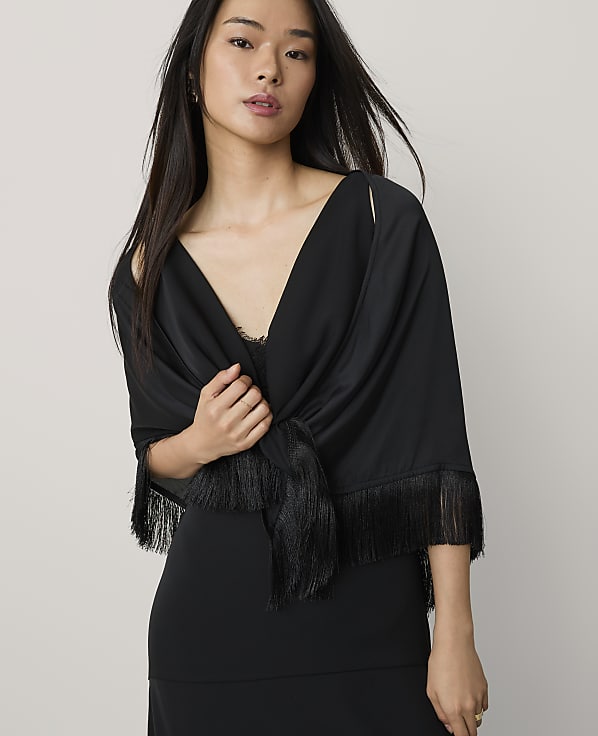 A woman wearing a black dress and a fringed black shawl stands against a plain, light-colored background.