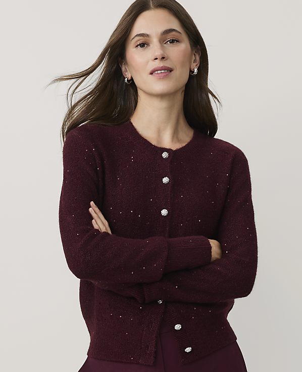 A woman with long brown hair wearing a sparkling burgundy cardigan stands with her arms crossed against a plain light gray background.