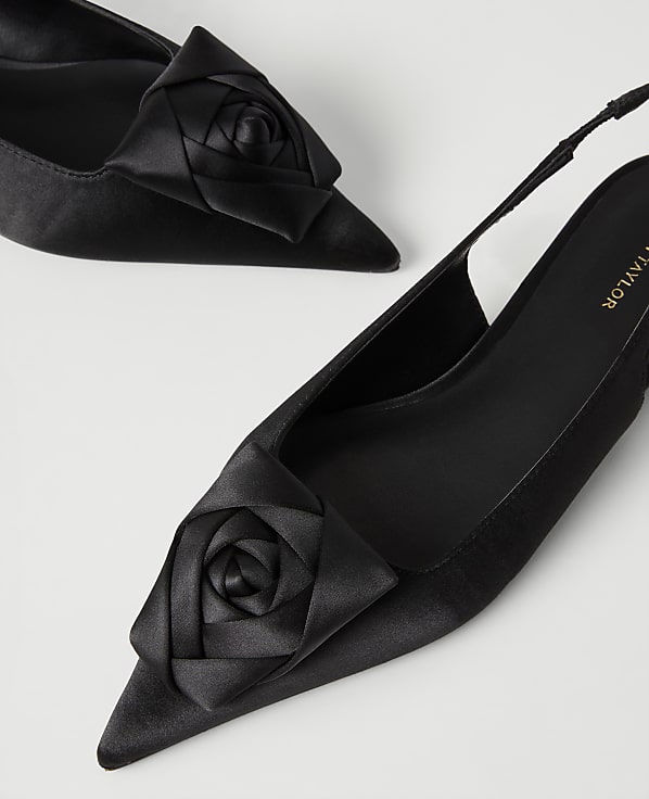 Two black satin pointed-toe heels with rose accents are positioned diagonally against a plain white background.