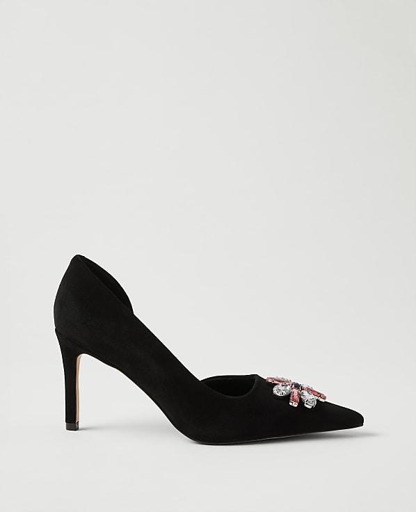 A black suede pointed-toe pump with a jeweled embellishment sits against a plain, light grey background.