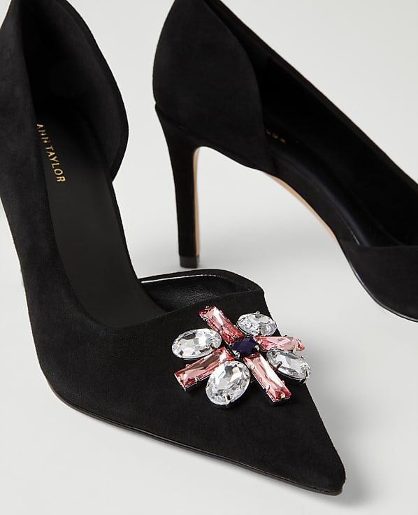 A pair of black suede pointed-toe pumps with a jeweled embellishment on the front are set against a plain white background.