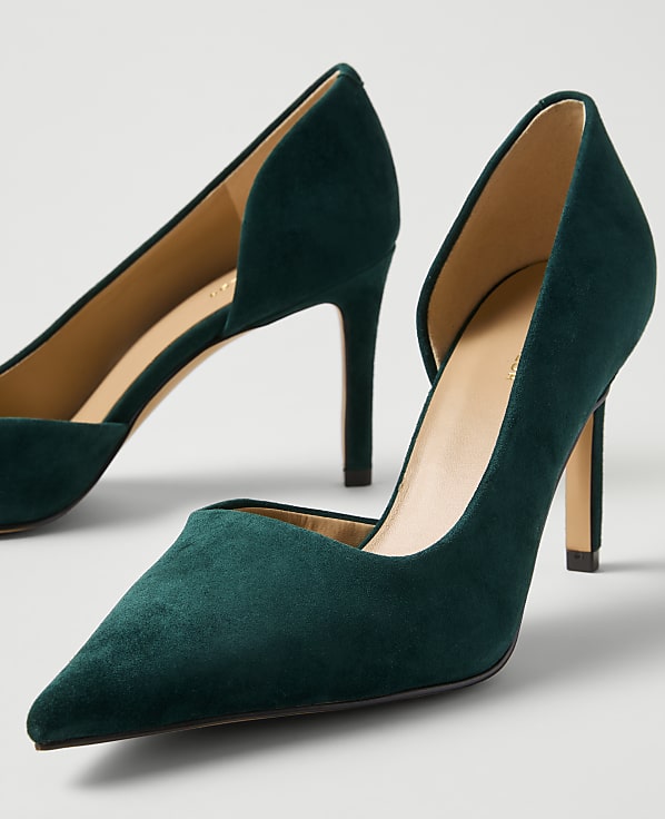 A pair of dark green suede pointed-toe pumps sit against a plain light gray background.