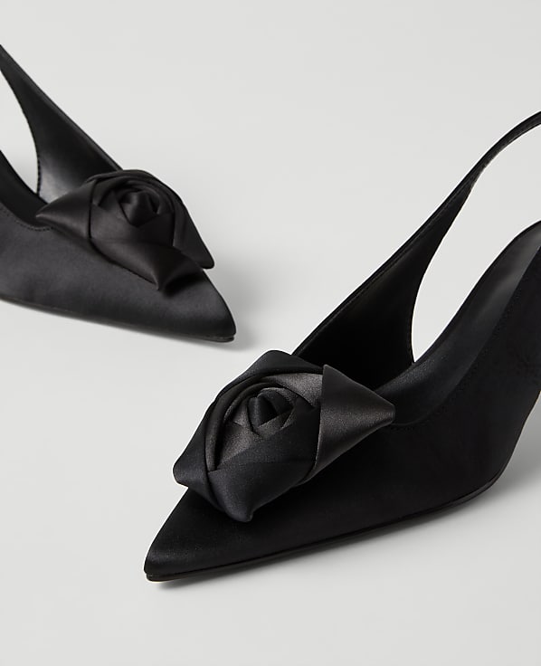 A pair of black satin slingback heels with rose-shaped ornaments on the toes are set against a plain light gray background.