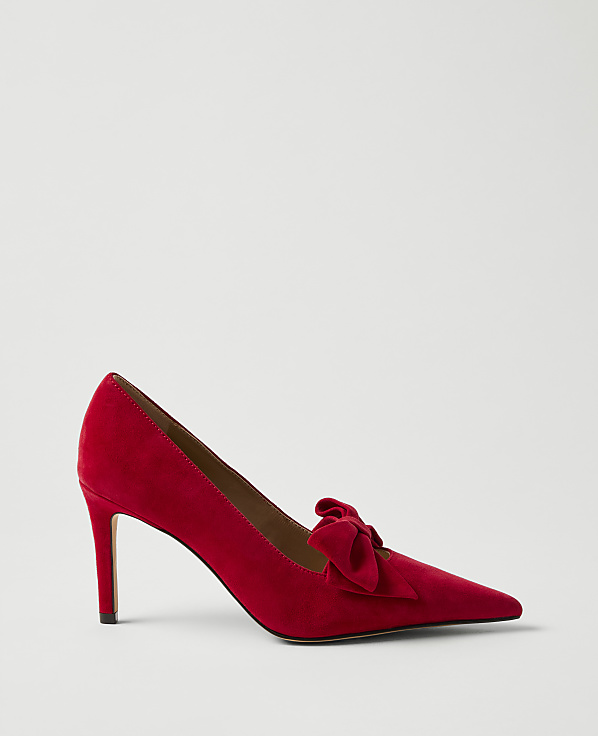 A red suede pointed-toe pump with a bow detail sits against a plain light gray background.