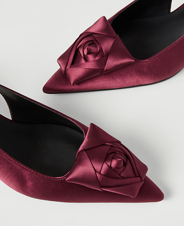 Two pointed-toe burgundy satin pumps with rose-shaped ornaments are positioned on a plain white background.