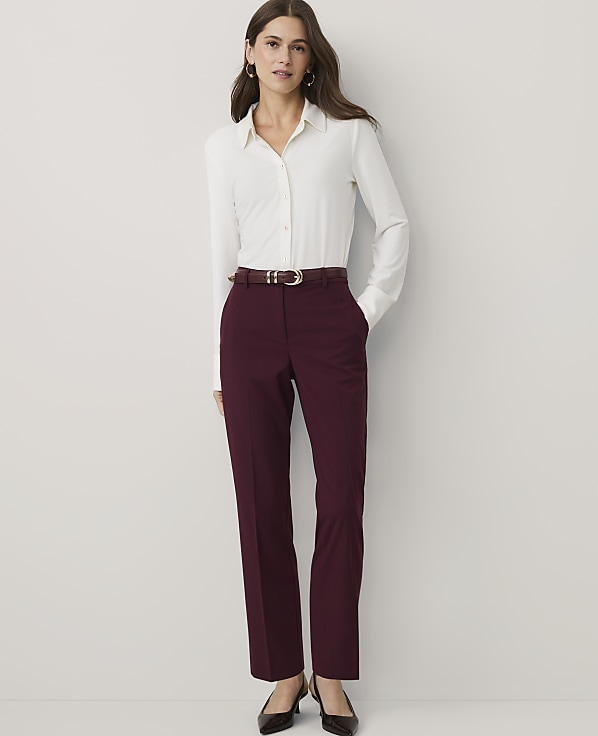 A woman wearing a white button-down shirt and burgundy trousers stands against a plain white background.