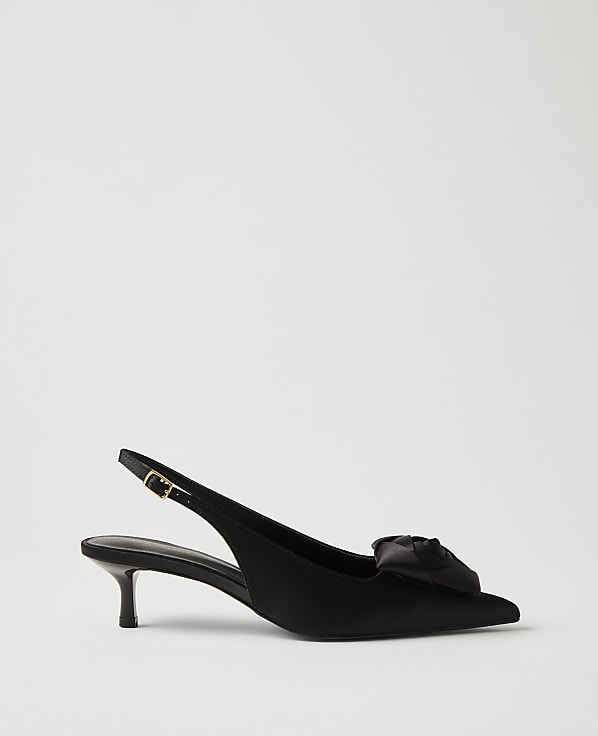 A black satin slingback heel with a decorative bow sits in the foreground against a plain, light gray background.