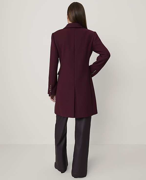 A woman stands with her back to the camera wearing a burgundy coat and dark wide-leg trousers against a plain light grey background.