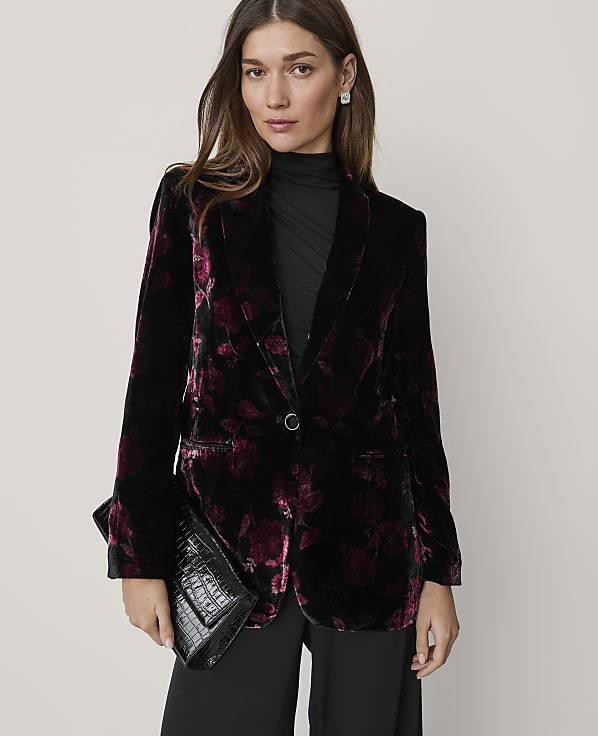 A woman wearing a floral velvet blazer and black clothing holds a black clutch against a plain, light-colored background.