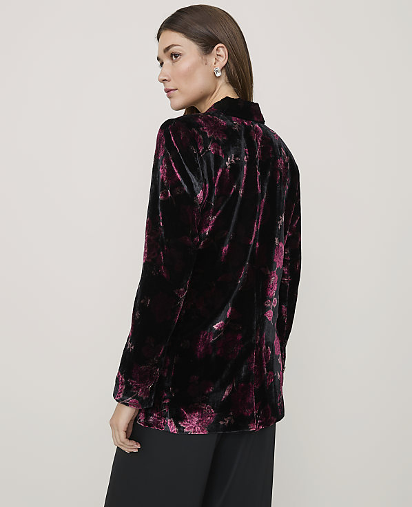 A woman wearing a black and purple floral velvet blazer and black trousers stands against a plain light gray background.