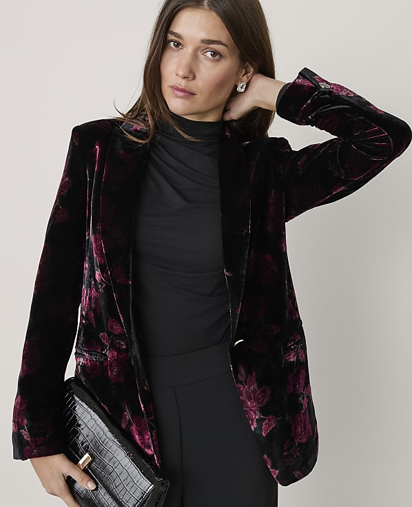 A woman wearing a floral velvet blazer and black clothing poses against a plain, light-colored background.
