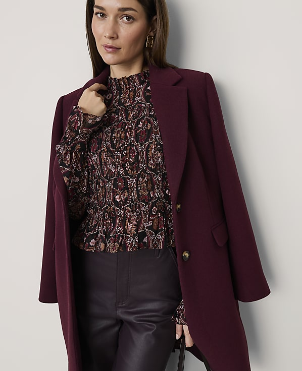 A woman wearing a patterned blouse, dark leather trousers, and a burgundy coat stands against a plain white background.