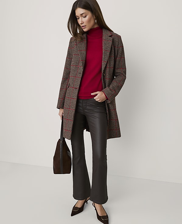 A woman wearing a plaid coat, red turtleneck, and dark faux-leather trousers stands against a plain, light-colored background.