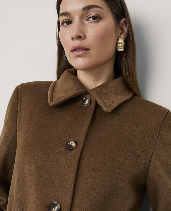 A woman wearing a brown wool coat and gold earrings poses against a plain, light-colored wall.