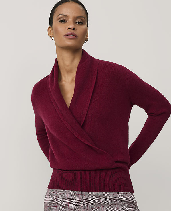 A woman wearing a deep red wrap sweater and plaid trousers stands against a plain light gray background.