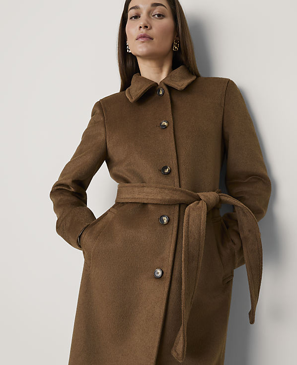 A woman wearing a brown belted coat stands against a plain white wall.