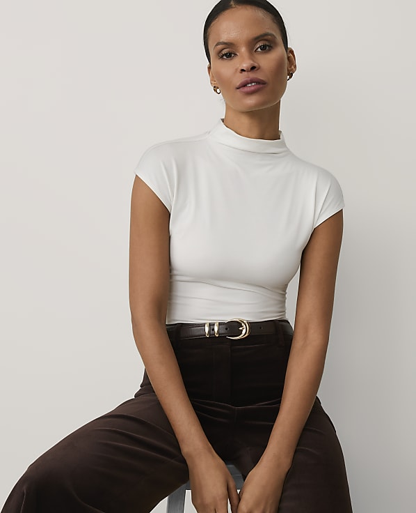 A woman wearing a white high-neck top and brown trousers sits against a plain, light-colored background.