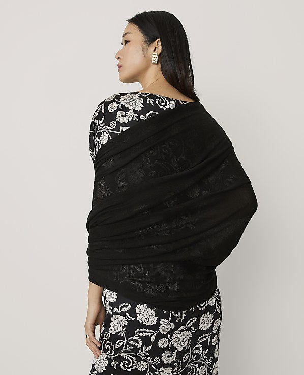 A woman wearing a black and white floral dress and a black shawl stands against a plain light gray background.