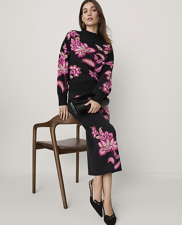 A woman wearing a black knit set with pink floral embroidery stands beside a wooden chair against a plain light grey background.
