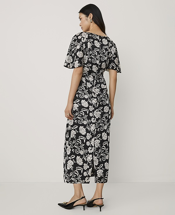 A woman wearing a black and white floral midi dress and black heels stands against a plain white background.
