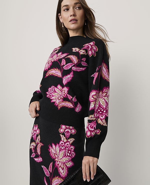 A woman wearing a black floral knit set stands against a plain, light-colored background.