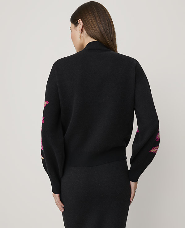 A woman stands with her back to the camera wearing a black sweater with pink floral accents and a black skirt against a plain light grey background.