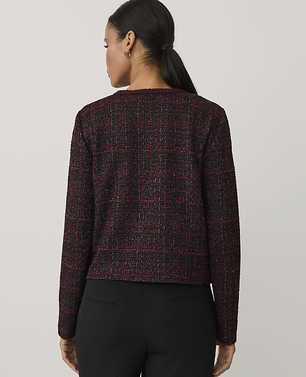 A woman wearing a red and black tweed jacket and black trousers stands with her back to the camera against a plain light grey background.
