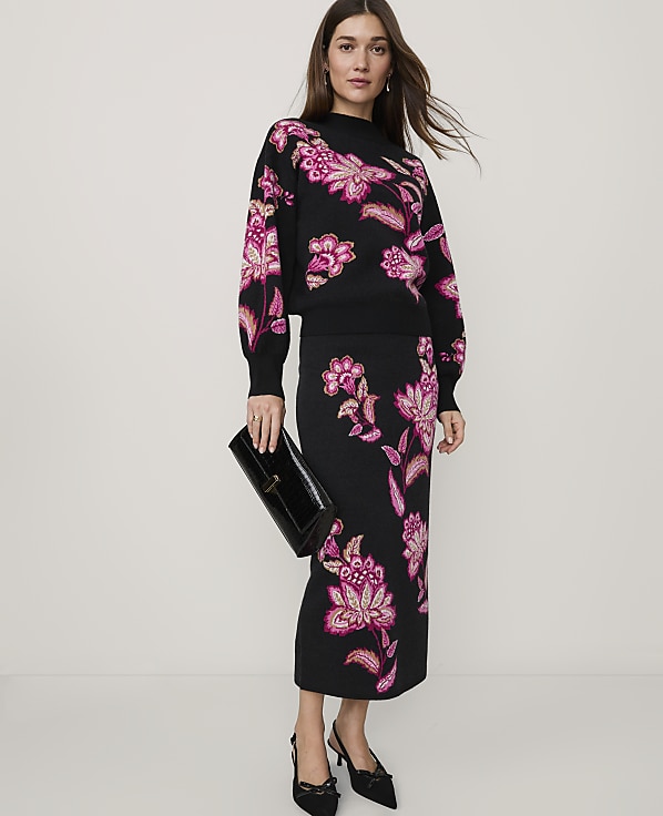 A woman wearing a black floral knit set and black heels stands against a plain, light-colored background.