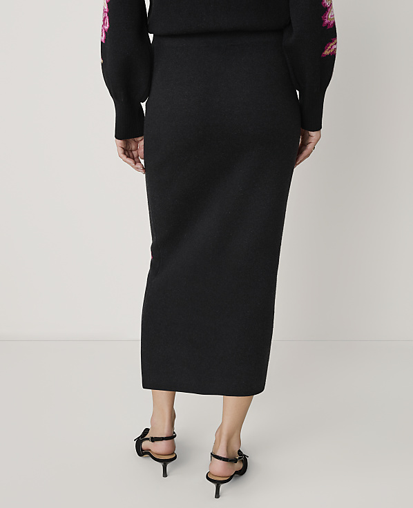 A person stands with their back to the camera wearing a black long-sleeved top with pink floral embroidery and a matching black midi skirt, set against a plain light gray background.