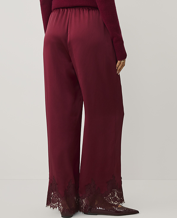 A person wears wide-leg burgundy satin trousers with lace hems and a matching sweater against a plain light gray background.