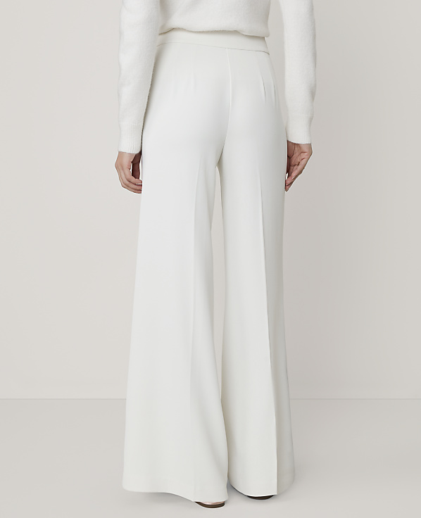 A person wearing a white sweater and wide-leg white trousers stands against a plain, light-colored background.