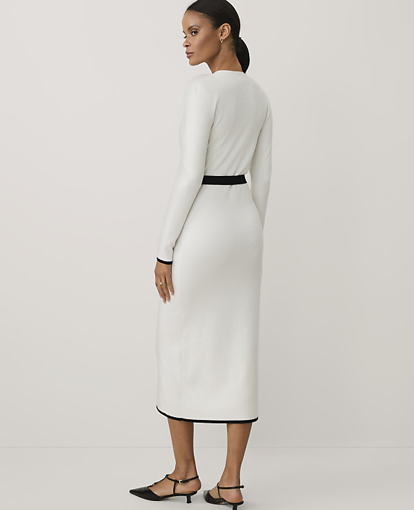 A woman wearing a long-sleeved white dress with black trim and black heels stands against a plain white background.