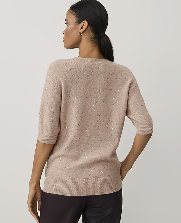A woman wearing a shimmering beige sweater and dark trousers stands with her back to the camera against a plain light gray background.