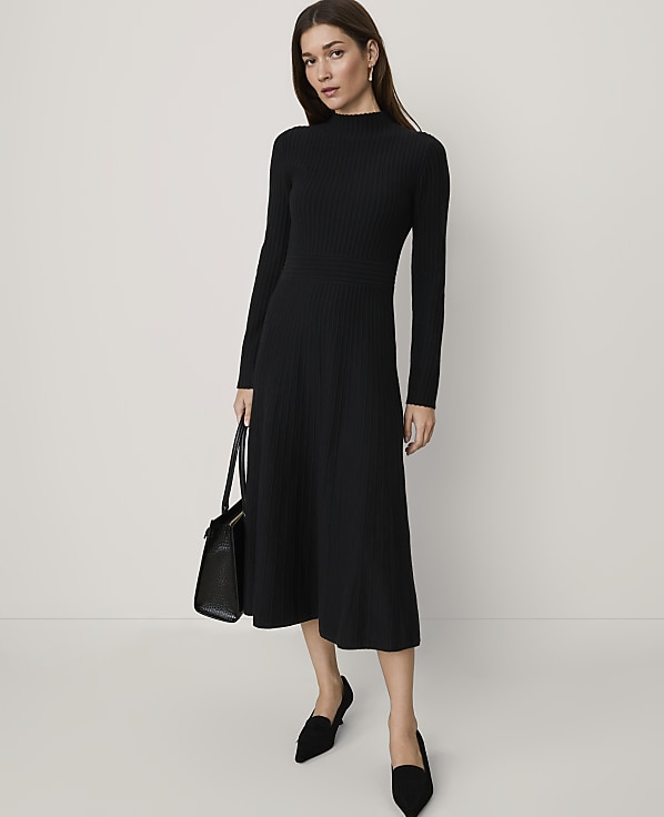 A woman wearing a long-sleeved black ribbed midi dress and black pointed-toe flats stands against a plain, light-colored background.