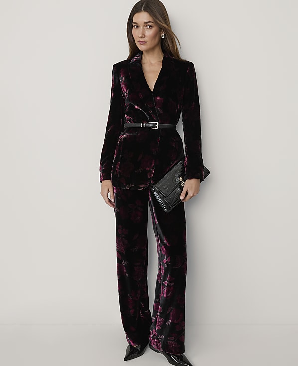 A woman wearing a dark purple velvet floral suit and holding a black clutch stands against a plain white background.