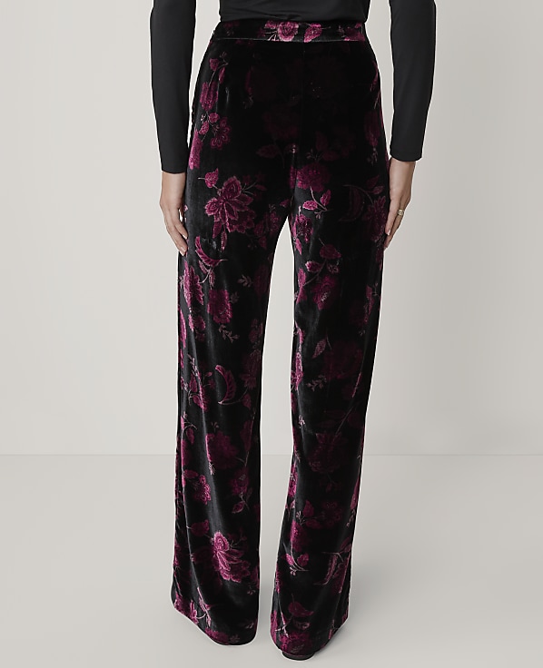 A person wears black long-sleeved top and wide-leg black velvet pants with a purple floral pattern against a plain light gray background.